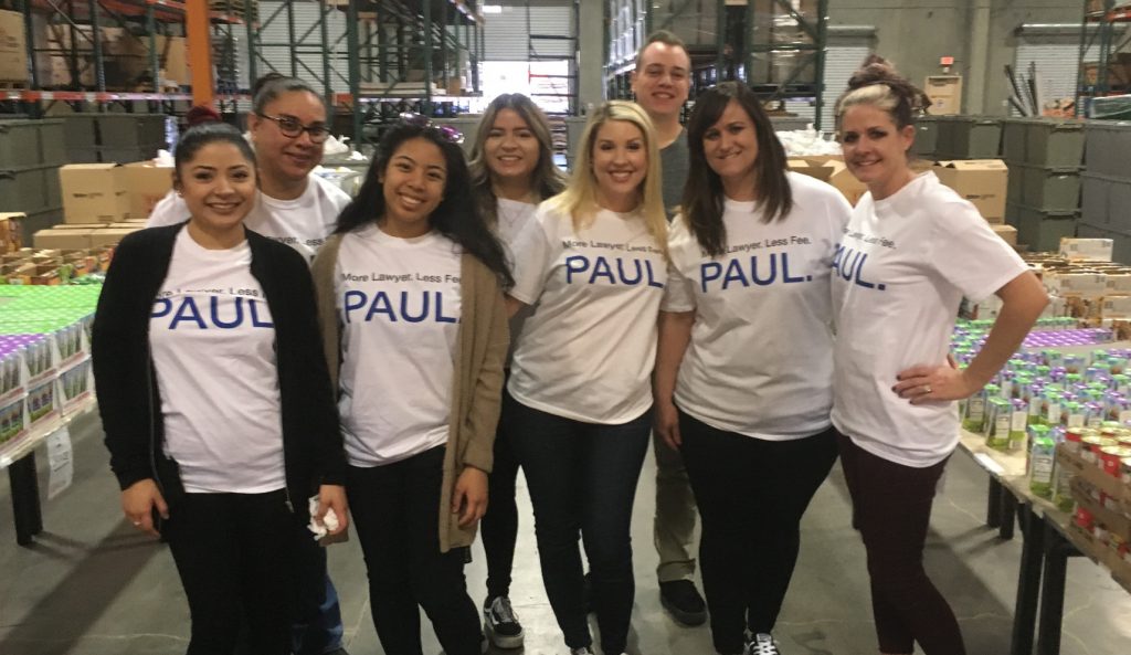 Community 1 A group of diverse smiling lawyers and staff from a Las Vegas personal injury law firm wearing branded T-shirts that say "More Lawyer, Less Fee, Paul" volunteer at a charity event in a warehouse setting, surrounded by stacked goods.