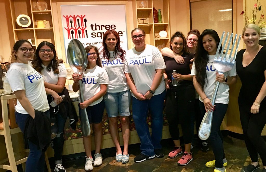 A group of diverse individuals, presumably from a Las Vegas personal injury law firm, poses with oversized kitchen utensils in front of a "three square" sign, indicating their volunteer work at a local food bank.