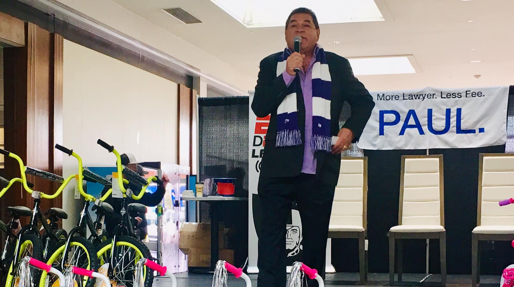 Las Vegas attorney Paul stands on a stage with a microphone, in front of a banner that says "More Lawyer. Less Fee. Paul.", and bicycles, likely at a community event.