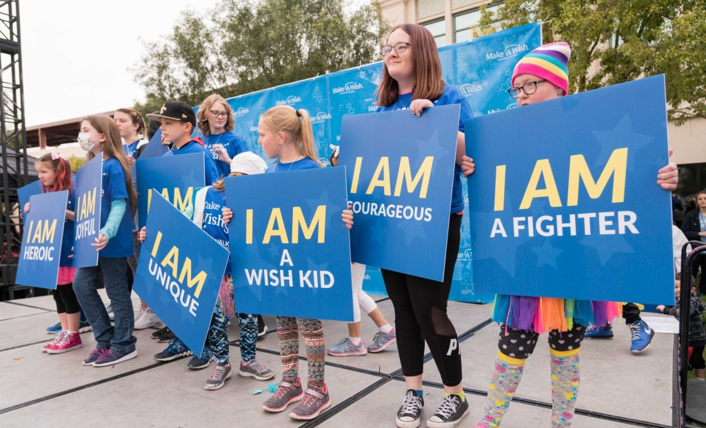 Community 3 A group of children stand on a stage holding signs with positive affirmations like "I AM a fighter," "I AM courageous," and "I AM unique," promoting a message of strength and resilience, possibly for a cause like Make-A-Wish, suitable for conveying a law firm's commitment to fighting for personal injury clients.