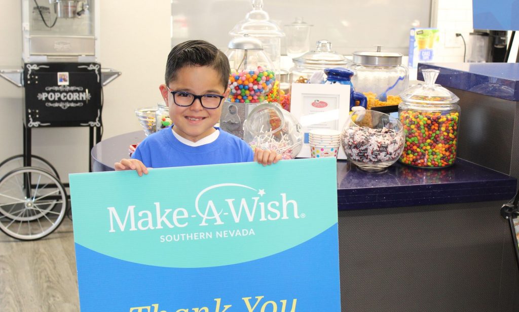 Smiling child holds a "Make-A-Wish Southern Nevada" sign in a Las Vegas law firm office, with jars of candy and a popcorn machine in the background, showcasing community involvement and support for children's causes related to injury.