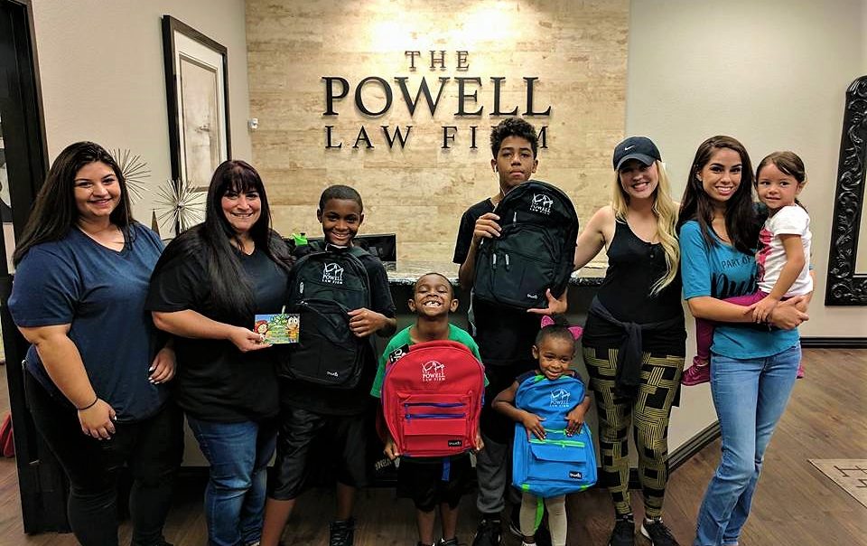 A group of children and adults pose with colorful backpacks featuring a law firm logo, standing in front of "The Powell Law Firm" sign, promoting community support and potentially appealing to families seeking personal injury legal services in Las Vegas.