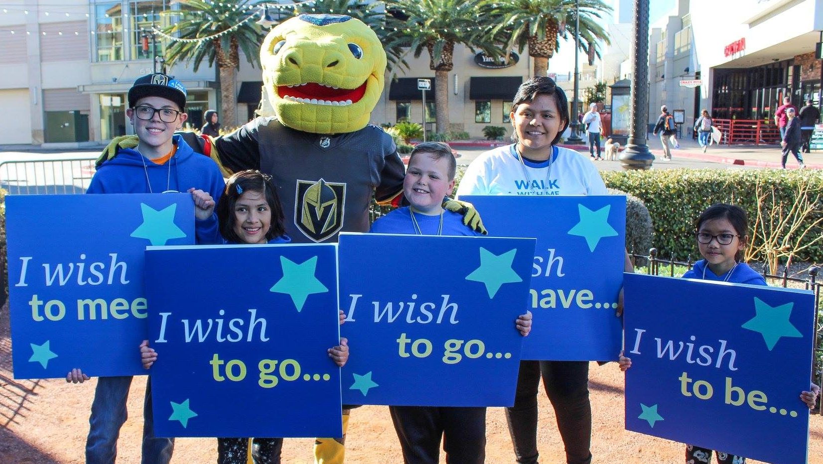A group of children stand holding signs with "I wish" phrases, accompanied by the Vegas Golden Knights mascot, Chance, at a charity walk in Las Vegas, potentially symbolizing hopes for recovery and support after personal injury cases.