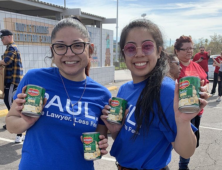 Smiling women holding cans of potatoes for a food drive, representing community support by a Las Vegas personal injury law firm.