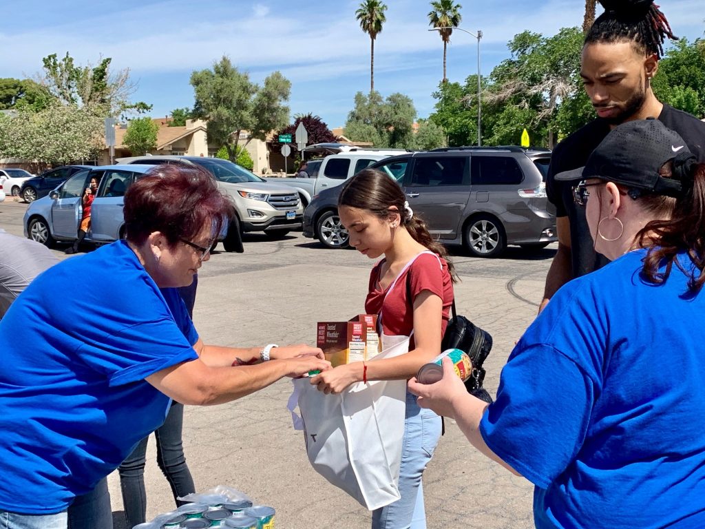 Community 7 A group of volunteers in blue shirts are assisting a young girl by placing food items into a white bag outdoors, with cars and palm trees visible in the background in Las Vegas.