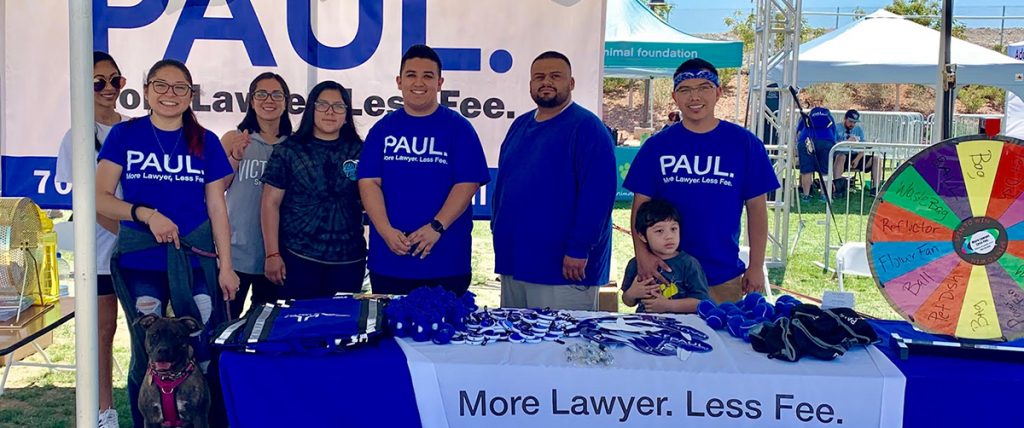Community 8 A group of people stand behind a table promoting a personal injury law firm in Las Vegas, with a wheel featuring prizes like "waste bag" and "pet dish", suggesting a community event possibly related to animal welfare.