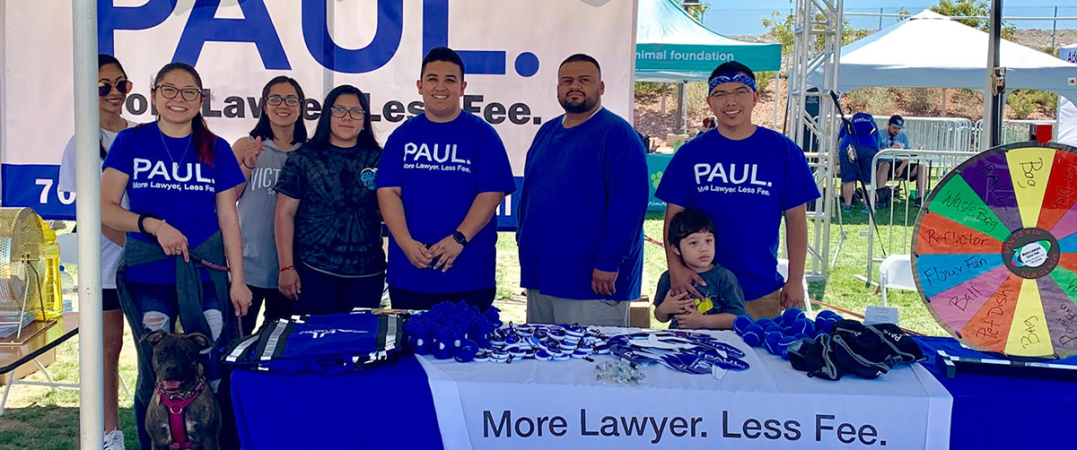 A group of people stand behind a table promoting a personal injury law firm in Las Vegas, with a wheel featuring prizes like "waste bag" and "pet dish", suggesting a community event possibly related to animal welfare.