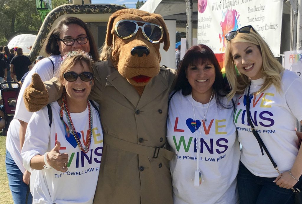 Group photo of smiling women and a dog mascot wearing "Love Wins" shirts for The Powell Law Firm, showcasing their support for the LGBTQ+ community and their Las Vegas legal services, including personal injury law.