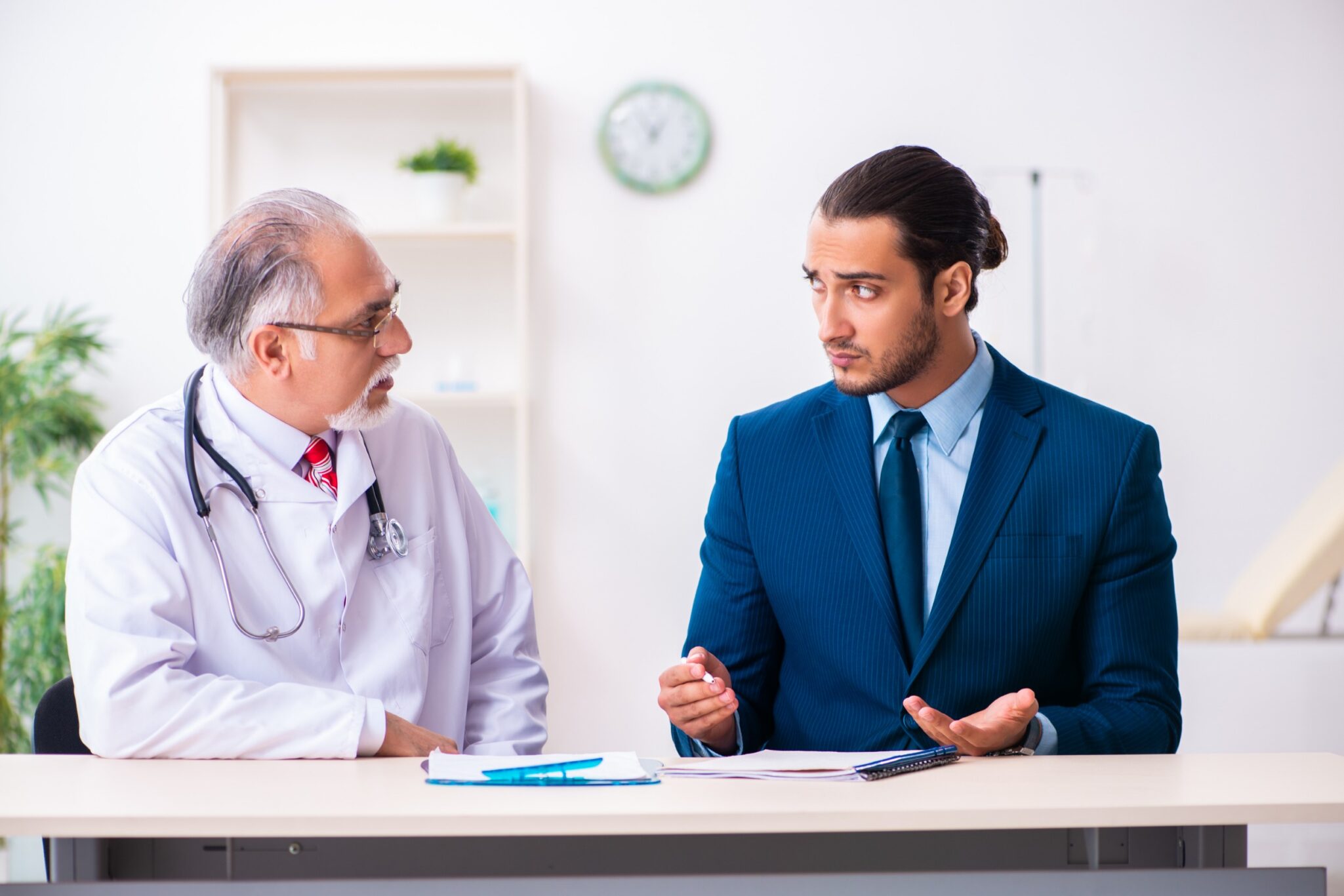 A doctor in a white coat with a stethoscope meets with a concerned man in a blue suit at a table, possibly discussing a personal injury case in a bright, clean office setting.