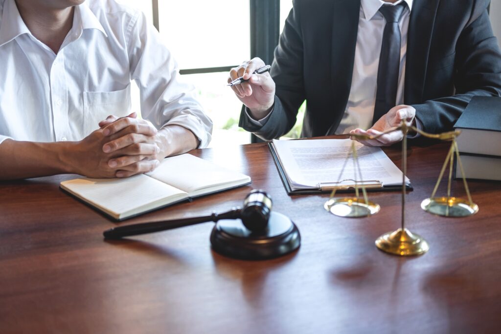 What Does More Lawyer, Less Fee Really Mean? 3 Image shows a lawyer discussing legal documents with a client at a wooden desk, with a gavel and scales of justice symbolizing law and fairness in personal injury cases.