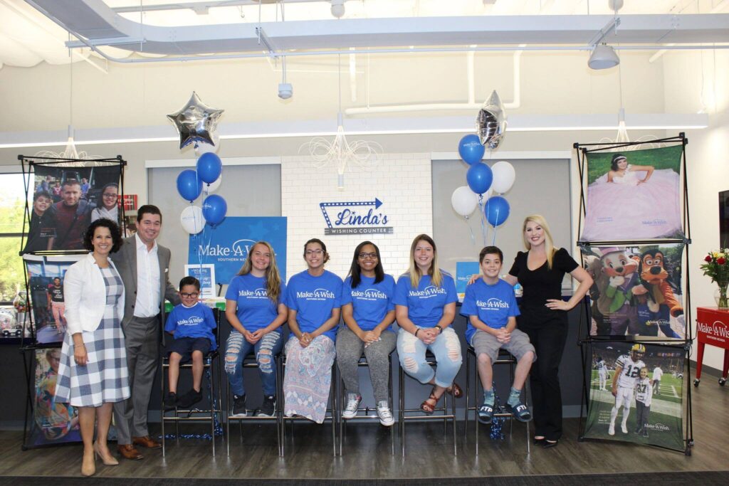 A group of people stand in front of a Make-A-Wish display inside Linda's Wishing Counter, including attorneys and children wearing Make-A-Wish t-shirts, symbolizing support for children's wishes and personal injury advocacy in Las Vegas.
