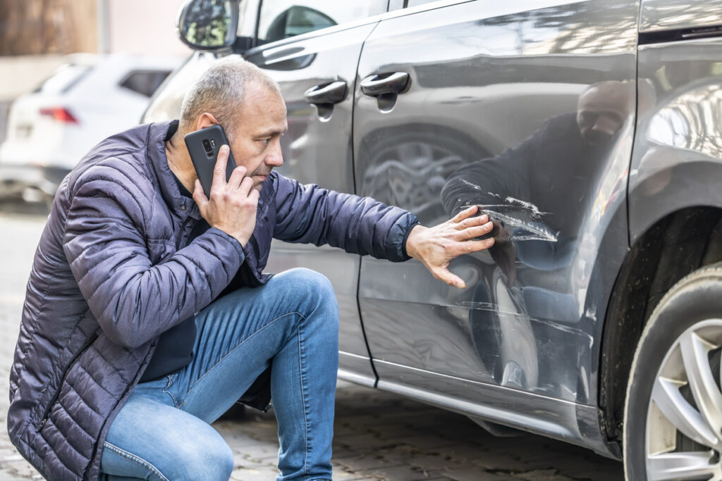 What Should You Do After an Accident? 2 A man kneels next to a gray car, examining damage from a car accident and talking on his cell phone, suggesting a call to a Las Vegas personal injury law firm for assistance.