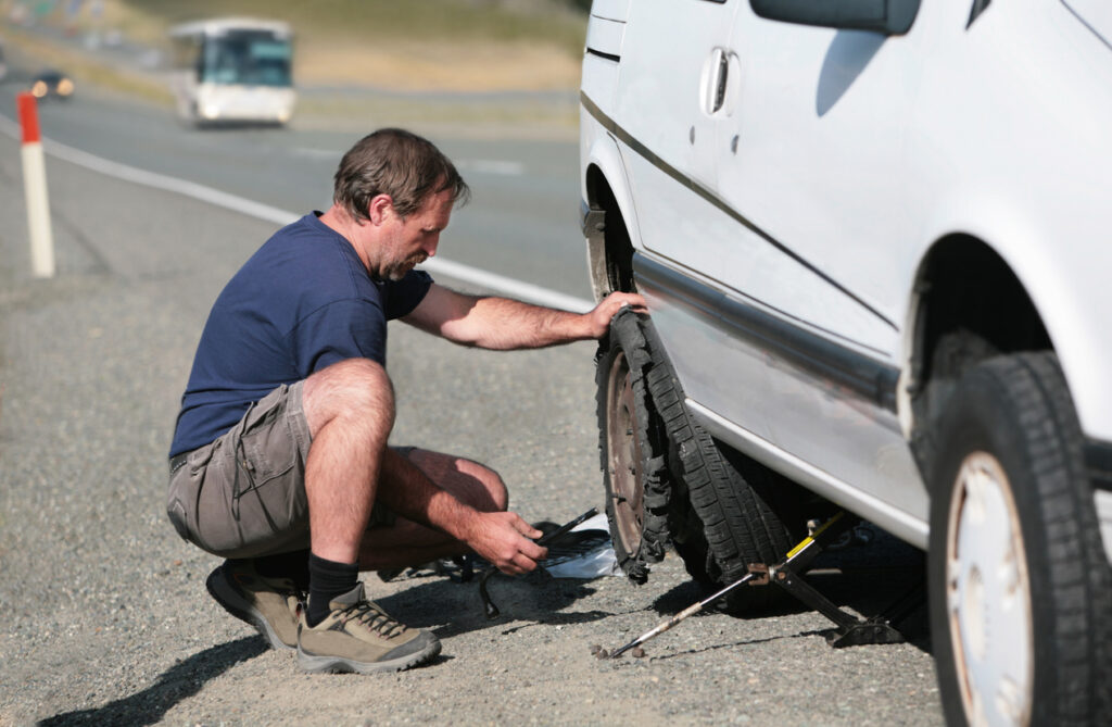 A Defective Tire Blowout Will Blow Up Your Life 1 Man changing a flat tire on the side of a road, highlighting the risks of roadside accidents and the need for personal injury legal assistance in Las Vegas.