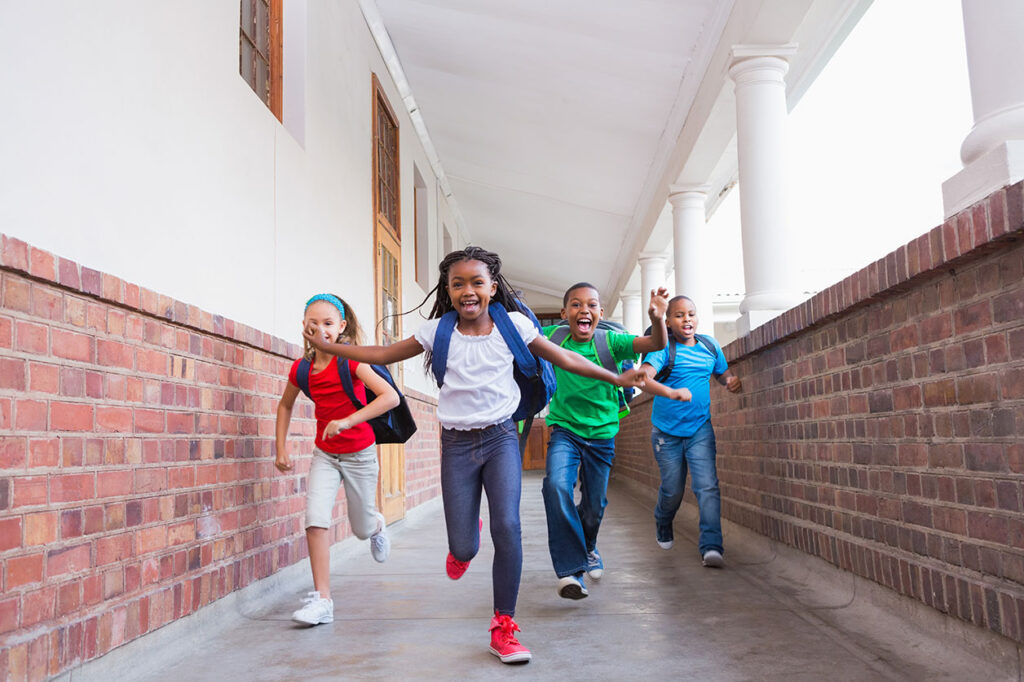 Four happy schoolchildren with backpacks are running outside a school building, potentially representing child injury risks and the need for legal representation from a personal injury law firm in Las Vegas.