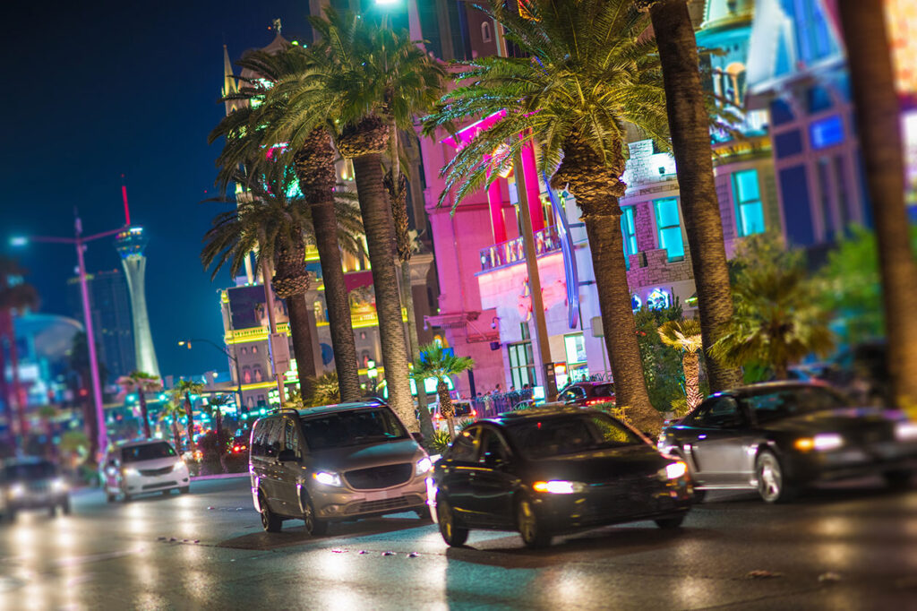 Night scene of cars driving on a Las Vegas street with bright lights and palm trees, ideal for a personal injury law firm ad.