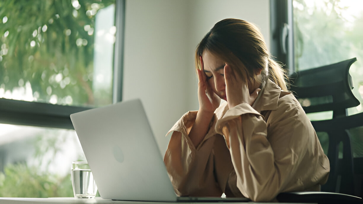 A woman holds her head in distress at a desk with a laptop, representing the stress and pain associated with personal injury in Las Vegas, prompting viewers to seek legal assistance.
