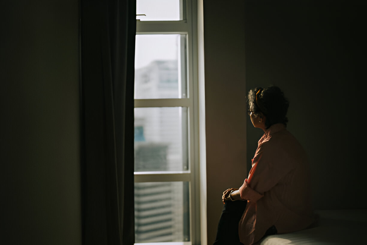 Lonely asian chinese senior woman standing in the dark behind the window looking